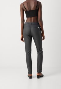 Frau steht barfuß in schwarzen, niedrighackigen Mules, trägt eng anliegende graue Hosen und ein schwarzes, bauchfreies Tanktop, blickt vor einer schlichten Hintergrundfläche nach hinten.