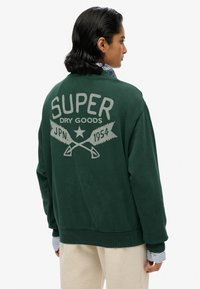 Felpa verde con una grande grafica grigia recante la scritta "SUPER DRY GOODS" e seghe incrociate sul retro. Materiale morbido e design comodo.
