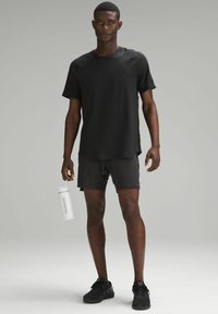Schwarzes, kurzärmeliges Sport-T-Shirt, kombiniert mit schwarzen Shorts, hält eine weiße Wasserflasche mit "lululemon"-Logo. Die Schuhe sind schwarz.
