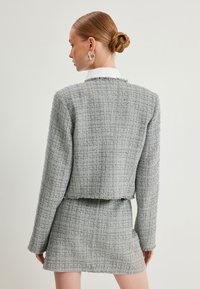 Harmaa tweed-lyhyt takki fransseilla reunoilla, yhdistettynä samasta kankaasta valmistettuun mini-hameeseen. Kankaan pinnassa on teksturoitu ruutukuvio.
