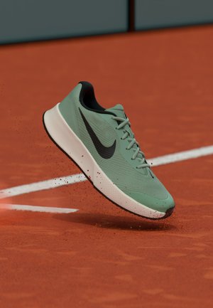 Roheline Nike jooksujalats õhus punase savitennisekatte kohal, valgete väljakupiiride lähedal laiali pillutatud saviosakestega.