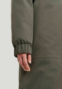 Oliven grøn jakkeærme med en tapered manschette, glat tekstur og en synlig søm langs armen, med en hånd hvilende ved siden af.
