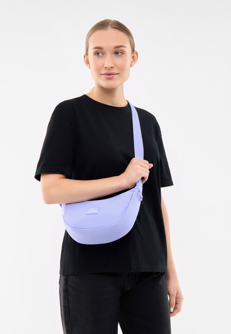Borsa a spalla in tessuto di un delicato viola, dalla forma curvata, con tracolla regolabile e un piccolo patch con logo; indossata su una camicia nera.