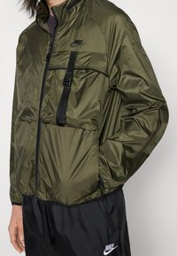 Olivgrön Nike-jacka med dragkedja framtill, svarta detaljer, elastiska muddar och en framficka som är säkrad med justerbar rem och sp änne.