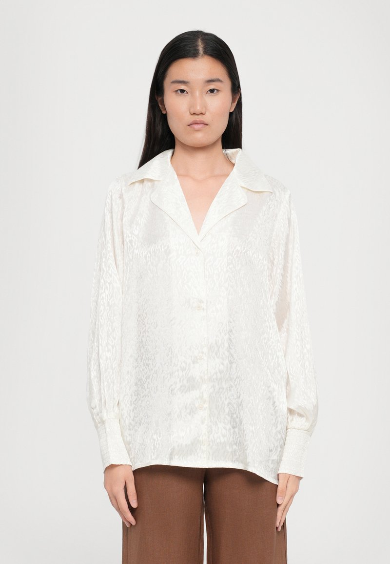 White satin shirt with a subtle pattern, large collar, and button-down front. Long sleeves with textured cuffs. Paired with brown trousers.