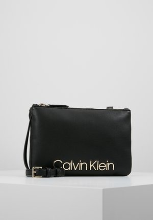 Czarna skórzana kopertówka Calvin Klein z złotym logo, zamkiem błyskawicznym i odpinanym paskiem, prezentowana na białym piedestale na szarym tle.