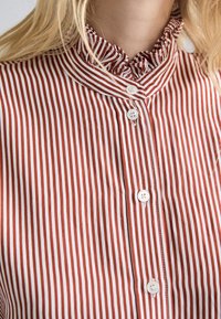 Primo piano di una blusa a righe verticali rossa e bianca con bottoni bianchi e un colletto alto arricciato su una persona con capelli biondi.