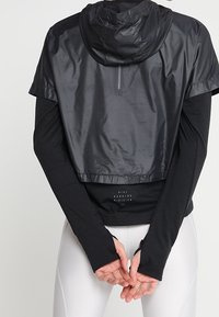 Veste de sport noire avec une couche supérieure à capuche courte et brillante et des manches longues ajustées en dessous. Comprend des passages pour les pouces et un logo au dos.