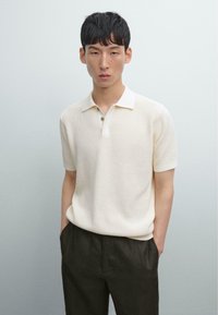 Polo a maniche corte color crema con maglia strutturata, pattina con due bottoni e colletto leggero, abbinato a pantaloni scuri.