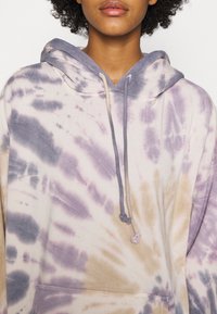 Felpa con cappuccio oversize in morbido cotone, caratterizzata da motivi viola, beige e grigi, con cappuccio regolabile e tasca a marsupio frontale.