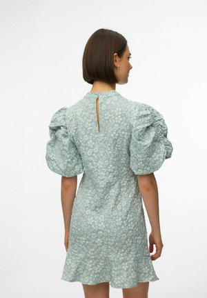 Donna con capelli corti castani che indossa un vestito verde chiaro a fiori con maniche a sbuffo e una piccola apertura a goccia sulla schiena.