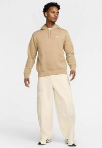 Felpa beige con tasca frontale e cappuccio regolabile, abbinata a pantaloni larghi color crema con tasche multiple e calzature con suola curva.