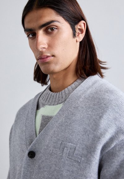 Cardigan lavorato a maglia di un grigio chiaro con scollo a V, dotato di un singolo bottone nero e dettagli del logo ricamato sul petto. Tessuto testurizzato.