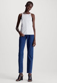 Hvit ermelaust topp med et teksturert mønster, kombinert med mørkeblå rette jeans og svarte hæl sandaler. Enkel, moderne design.