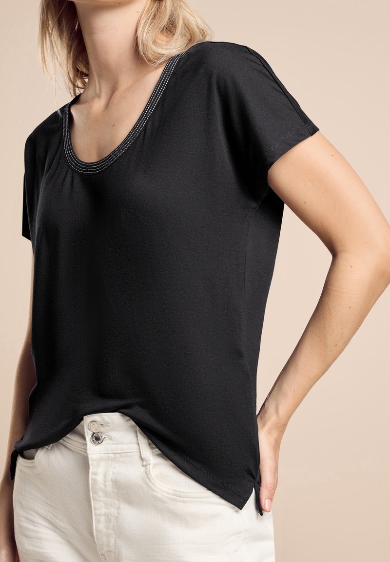 Svart kortärmad topp med vid halsringning och kontrasterande sömmar, med en avslappnad passform och sidoklyftor. Bärs ihop med ljusfärgade byxor.