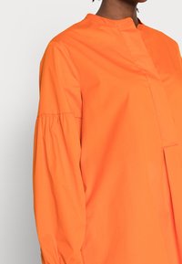 Chemisier en coton orange avec un col montant, des manches plissées et une coupe droite. Texture lisse avec un design minimaliste.