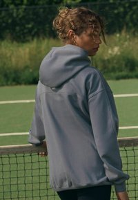 Grauer Hoodie mit weicher, strukturierter Oberfläche, lockerer Passform und Kordelzugkapuze. Verfügt über dezentes Branding auf der Rückseite in der Nähe des Kragens.