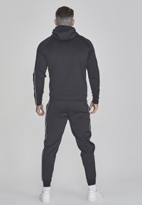 Schwarzes Sweatshirt- und Jogginganzug-Set mit Kapuze, ausgestattet mit weißen Seitenstreifen. Glatter Stoff und taillierte Passform. Weiße Sneaker runden den Look ab.