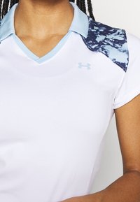 Valkoinen urheilupaita, jossa on vaaleansininen kaulus ja kuvioidut siniset yksityiskohdat hartioilla. Includes v-kaula-aukko ja Under Armour -logo.