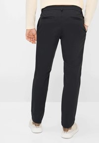 Pantalon noir à texture lisse, doté de deux poches arrière, conçu pour une coupe slim, associé à des baskets de couleur claire.