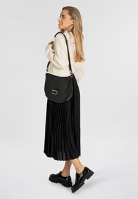 Borsa a spalla in pelle nera con finitura testurizzata, caratterizzata da accenti in metallo dorato e un design strutturato. Il modello indossa una gonna plissettata nera.