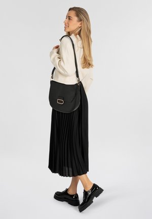 Borsa a spalla in pelle nera con finitura testurizzata, caratterizzata da accenti in metallo dorato e un design strutturato. Il modello indossa una gonna plissettata nera.