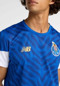 Blå atletisk trøje lavet af tekstureret stof. Har hvide skulderdetaljer, gyldt 'NB' logo og FC Porto-emblemet på brystet.