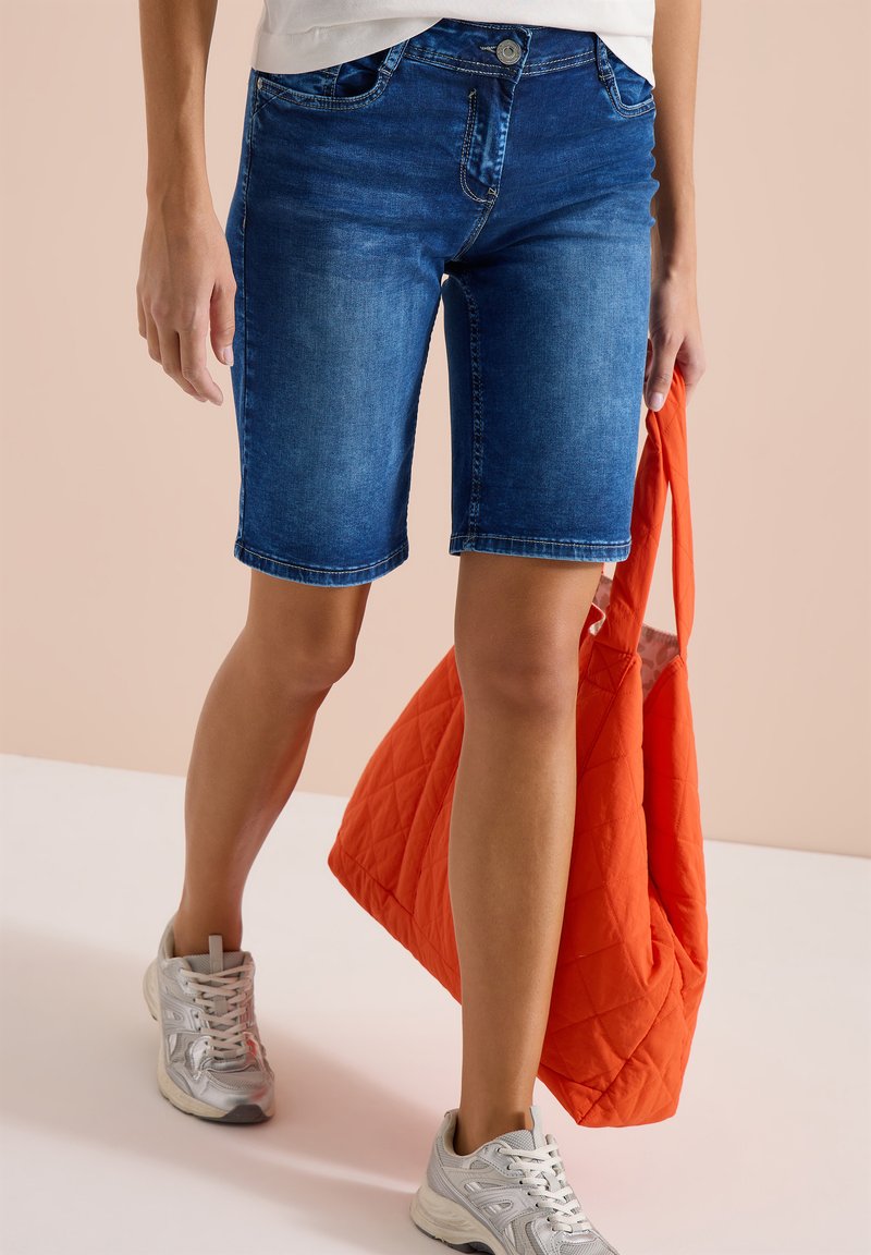 Personne portant un short en jean bleu au genou, des baskets blanches, tenant un grand sac cabas matelassé orange sur un fond beige.