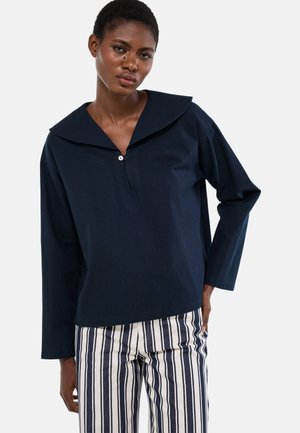 Femme portant un haut à manches longues bleu marine avec un col large et un pantalon à rayures verticales blanches et bleues, debout avec une main dans sa poche.