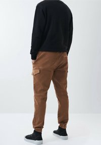 Bruna cargobyxor med elastiska manschetter, sidofickor och en avslappnad passform. Bärs med en svart långärmad tröja och svarta högskor.