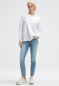 Blonde Frau steht da und trägt einen weißen Sweatshirt mit Text, hellblaue Jeans mit Aufschrift und weiße Sneakers, mit einer Hand in der Tasche.