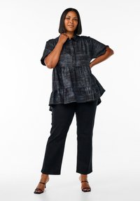 Woman stands confidently with short black hair, wearing a loose black textured blouse, black pants, and black heeled sandals on white background.