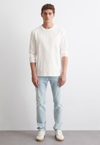 Langärmliges weißes Baumwoll-T-Shirt mit einer Brusttasche, kombiniert mit hellblauen Jeans und weißen Sneakers mit hellbraunen Sohlen.