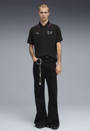 Giovane uomo che indossa una polo nera BMW Motorsport, pantaloni neri a gamba larga e scarpe nere, in piedi con una mano in tasca e cuffie che pendono.