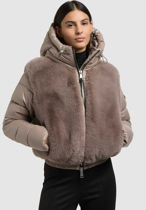 Femme portant une veste matelassée taupe avec une grande capuche et un devant en fourrure douce, mains dans les poches, debout devant un fond clair uni.