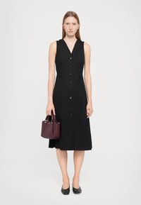 Robe noire sans manches avec un décolleté en V, dotée d'un devant à boutons et de deux poches latérales. Le modèle tient un petit sac à main bourgogne.