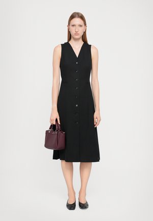 Schwarzes ärmelloses Kleid mit V-Ausschnitt, mit Knopfleiste vorne und zwei Seitentaschen. Das Modell hält eine kleine bordeauxfarbene Handtasche.