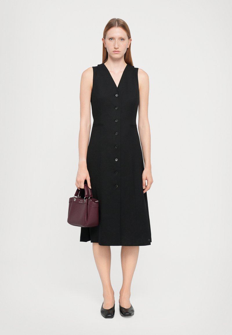 Robe noire sans manches avec un décolleté en V, dotée d'un devant à boutons et de deux poches latérales. Le modèle tient un petit sac à main bourgogne.