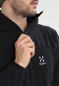 Veste softshell noire avec col montant, fermeture éclair à l'avant et logo sur la poitrine. Dotée d'un tissu texturé et d'une poche latérale discrète.