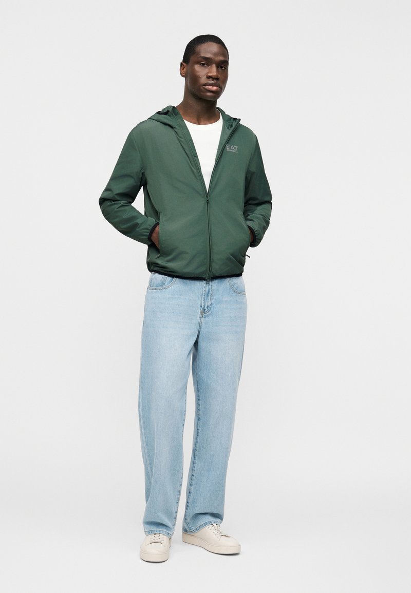 Άνδρας που στέκεται με τα χέρια στις τσέπες, φοράει πράσινο σακάκι με κουκούλα, λευκή μπλούζα, ανοιχτό μπλε τζιν και λευκά αθλητικά παπούτσια σε λευκό φόντο.