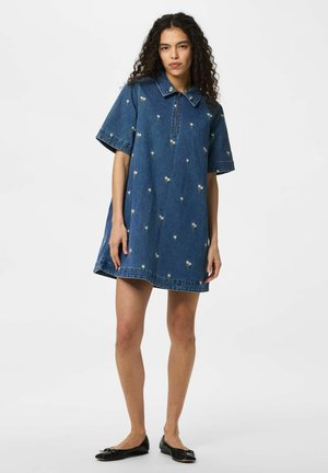 Femme aux cheveux bouclés portant une robe en denim bleu avec une petite broderie florale blanche et des ballerines noires, debout sur un fond blanc.