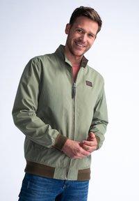 Zelená bomber bunda se zipem vpředu, žebrovanými manžetami a lemem. Obsahuje hnědou koženou nášivku a hladkou, lehkou texturu materiálu.