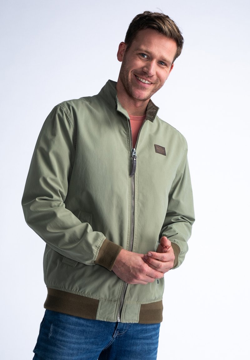 Zelená bomber bunda se zipem vpředu, žebrovanými manžetami a lemem. Obsahuje hnědou koženou nášivku a hladkou, lehkou texturu materiálu.