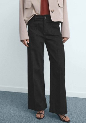 Person wearing black wide-leg pants, beige jacket, rust-colored top, and black strappy high-heeled sandals standing on gray carpet.
