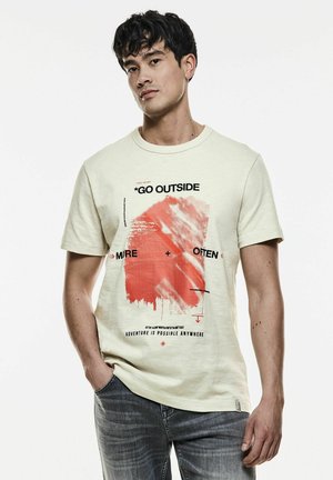 Camiseta de algodón amarillo claro con un diseño gráfico en rojo y el texto "SAL DE CASA" en negro, combinada con jeans grises.