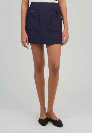 Femme portant une mini-jupe bleu marine avec deux poches avant à bouton, associée à des ballerines noires à bout pointu sur un fond clair uni.