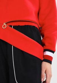 Sweat-shirt rouge avec une fermeture éclair dorée partiellement ouverte sur un pantalon noir avec un liseré blanc, et une main reposant sur le côté.