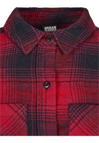 Chemise en flanelle à carreaux rouges et marine avec un col à boutons, des boutons noirs et une poche sur la poitrine. La texture du tissu semble douce et chaude.