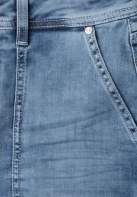 Tissu en denim bleu clair avec coutures visibles, une passants de ceinture et une poche avant sécurisée par un petit bouton à rivet métallique.