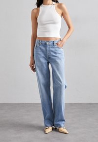 Jeans rectos de tiro alto en azul claro, con cierre de cremallera y botón, y dos bolsillos frontales. Combinados con una blusa sin mangas blanca.